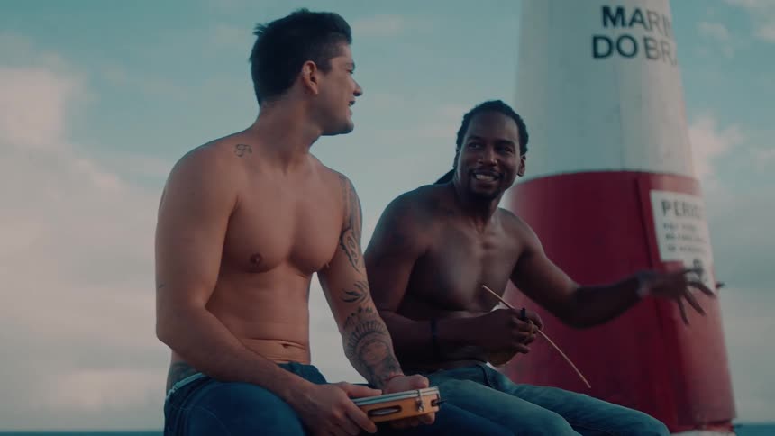 Two shirtless men sit near a lighthouse under a partly cloudy sky both holding musical instruments