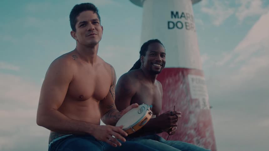 Two shirtless men sit near a redandwhite lighthouse while waves crash behind them