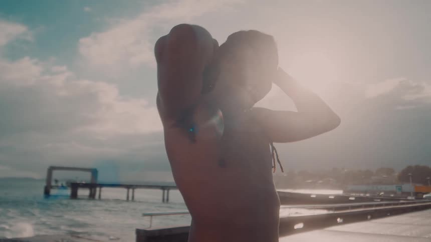 A bare chested man with dark hair smiles with his arms raised posing by a choppy ocean with a pier and buildings in the distance under a bright sky