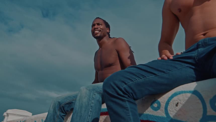 Two shirtless men sit outdoors near a building under a partly cloudy sky one with visible tattoos and the other with long hair