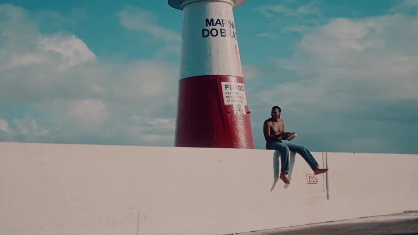 A shirtless man sits on a barricade with a lighthouse behind him