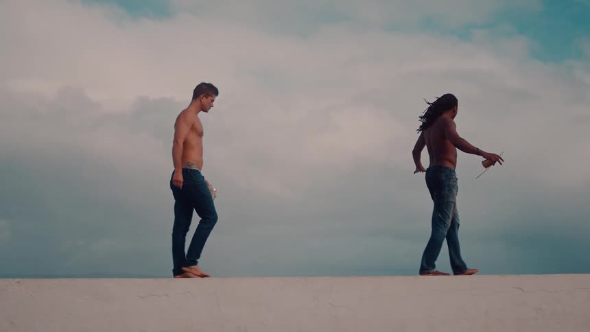 Two men walk on a concrete path toward a lighthouse at midday shirtless and wearing jeans