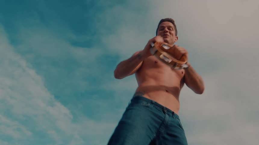 Shirtless man dancing with a tambourine under cloudy blue skies having fun