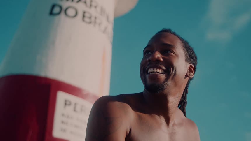 A shirtless man with a beard smiles broadly against a bright blue sky