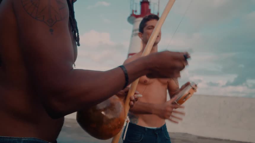 Two shirtless men are outdoors one playing a bell and the other a tambourine with a lighthouse in the background