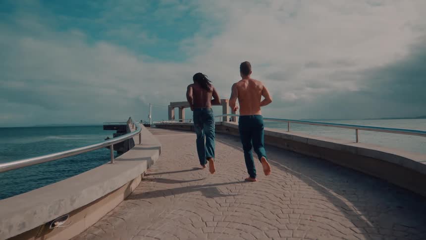 Two people jog barefoot along a curved oceanside path under a partly cloudy sky