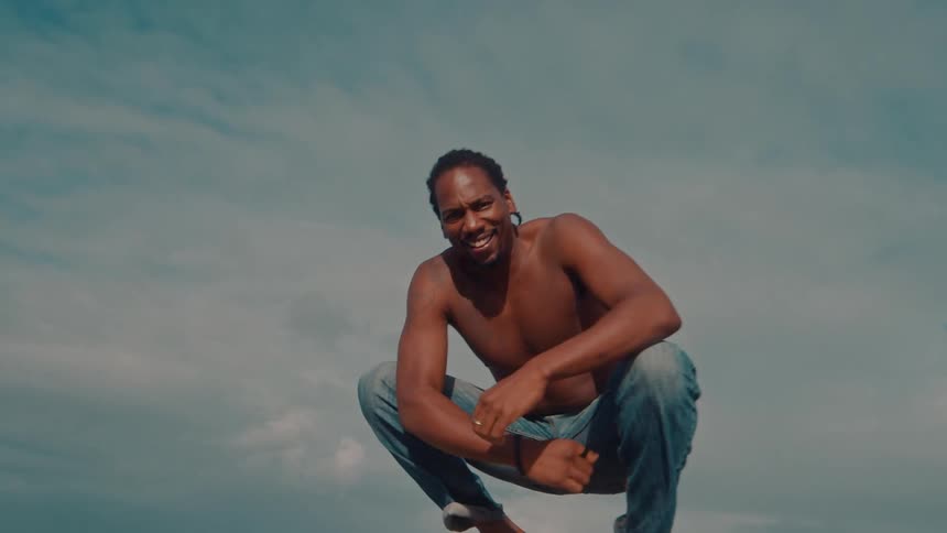 A man squatting at the beach shore gesturing with his hand to call someone