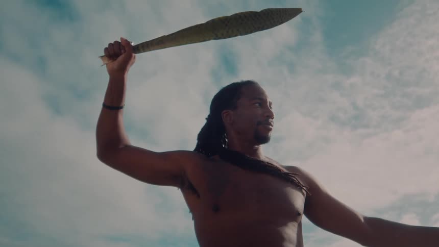 A pacific islander man holds a clubbed weapon over his head in the wind