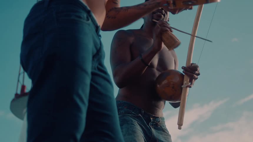 Two shirtless men stand on the coast around midday holding and playing musical instruments while singing a song