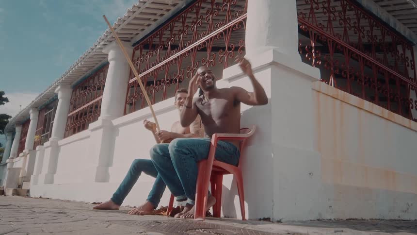 Man blowing a kiss with his hands while sitting next to someone else holding a berimbau