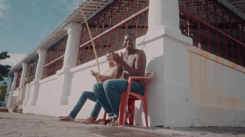 Two shirtless men sitting on a chair clapping and playing a berimbau