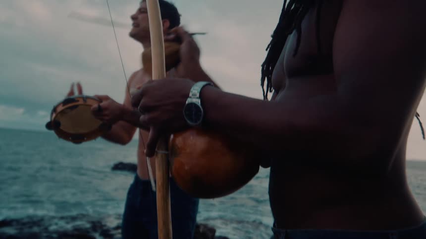 Two men are playing instruments on the rocks by the water