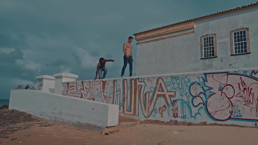 Two shirtless men walk along a graffiticovered wall with a white building and overcast sky in the background