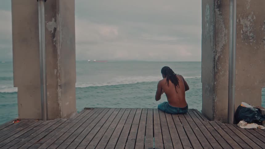A black man with dreads sits on the edge of the coast as ocean waves hit the shore around midday facing the ocean