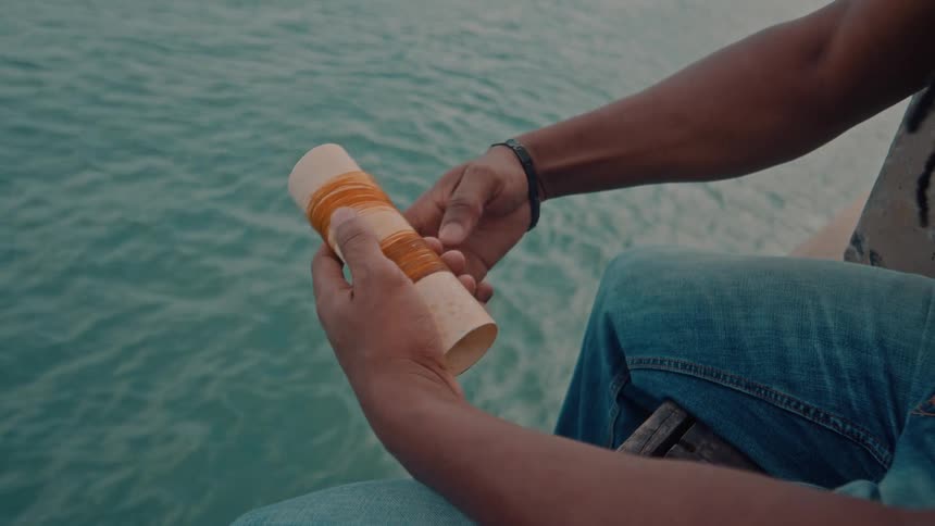 A man sits on a platform over the ocean tying a rope around a cylinder around midday