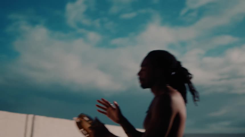 A shirtless person plays a tambourine with one hand casting a shadow on a sunlit wall