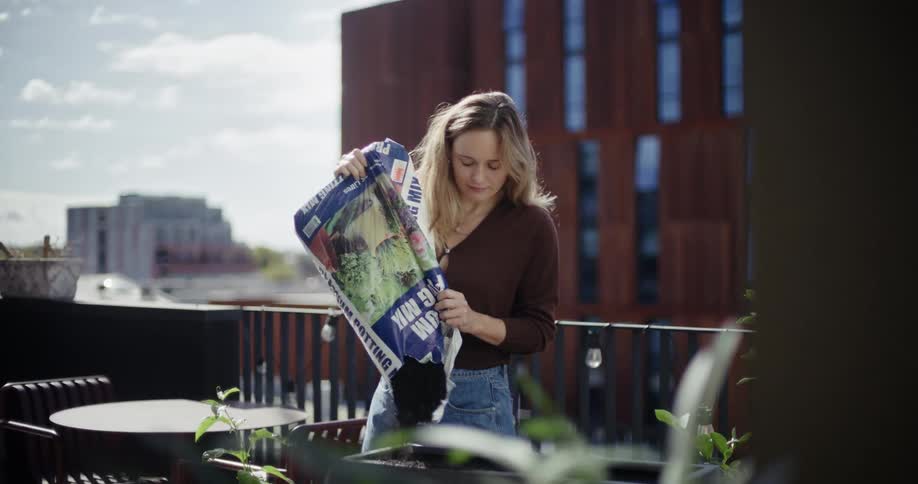 A woman on a balcony pours potting mix from a bag into planters against an urban backdrop