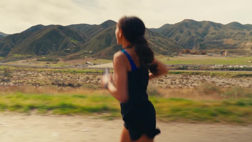 A young woman jogs along a scenic trail smiling back at the camera with mountains in the background