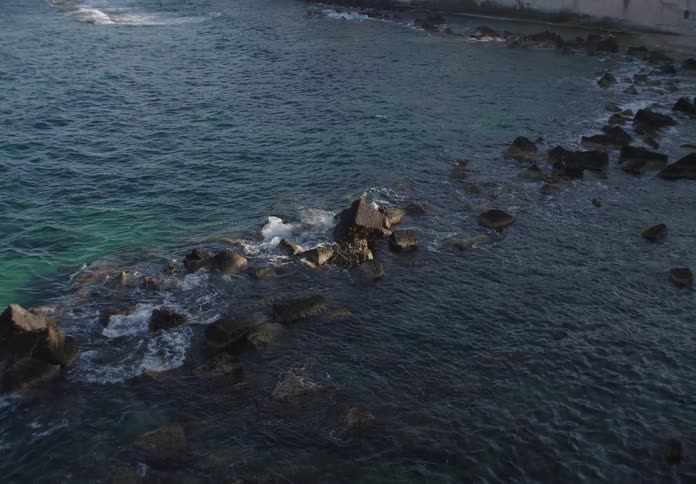 Waves crash gently against scattered rocks in shallow coastal waters near a shoreline