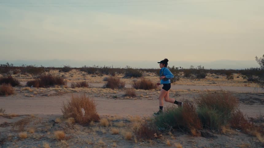 A woman is jogging through a dry desertlike landscape under a calm sky