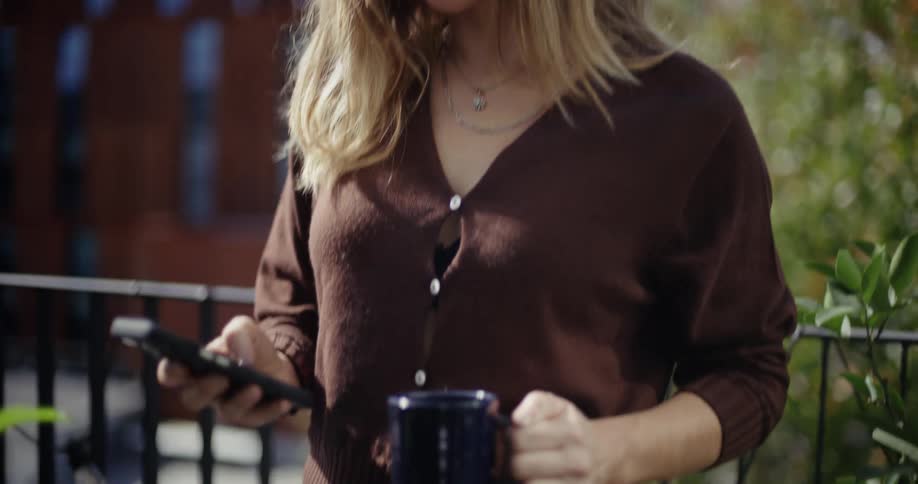 A woman stands outdoors holding a mug and phone smiling while texting