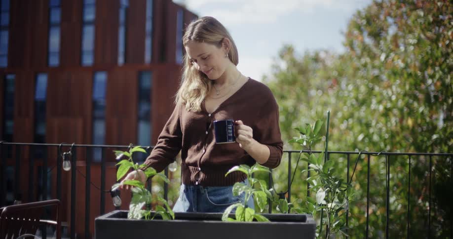 A woman tends to plants on a sunny balcony while holding a coffee mug