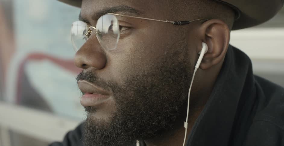 A man sits outside with his earbuds in and the road around him is reflected on his glasses