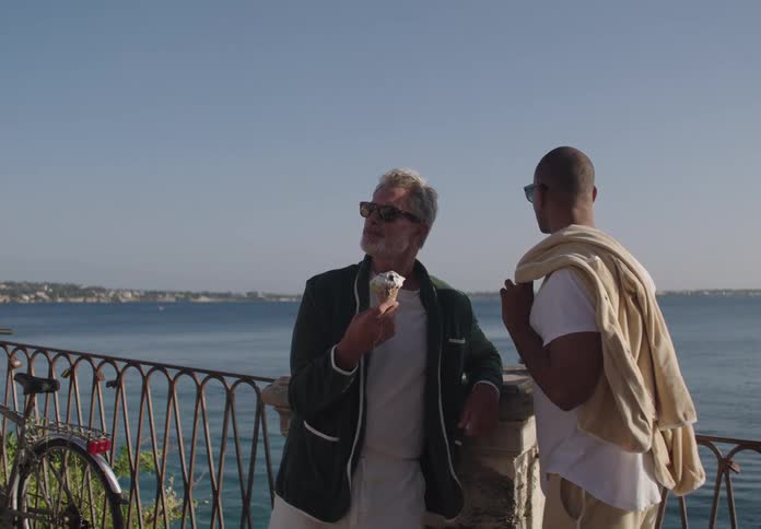 Two men relax by the seaside one eating ice cream