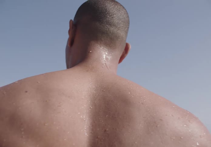 A young man bareback and drenched with water as if he just came out of a pool stands around midday