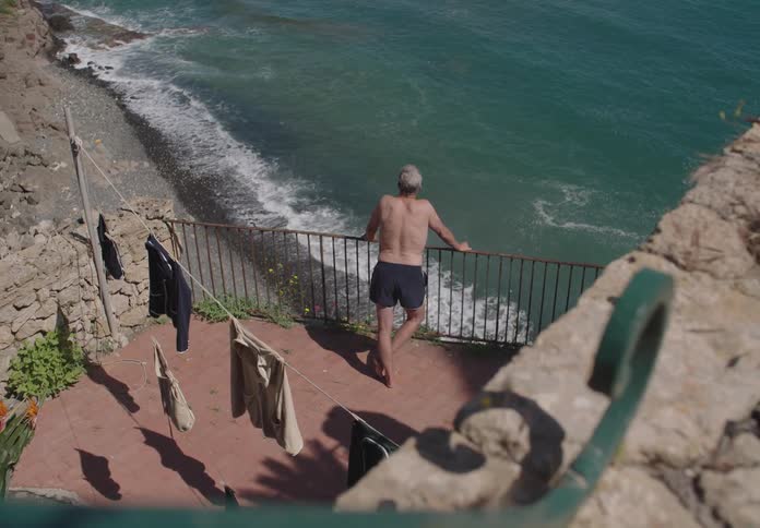 A shirtless man stands on a seaside terrace gazing out over the ocean near a stone wall