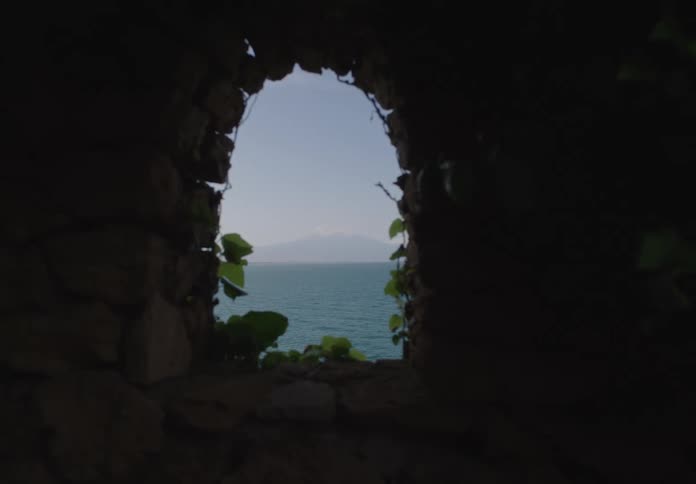 A coastal view is seen through a small stone window covered in vines
