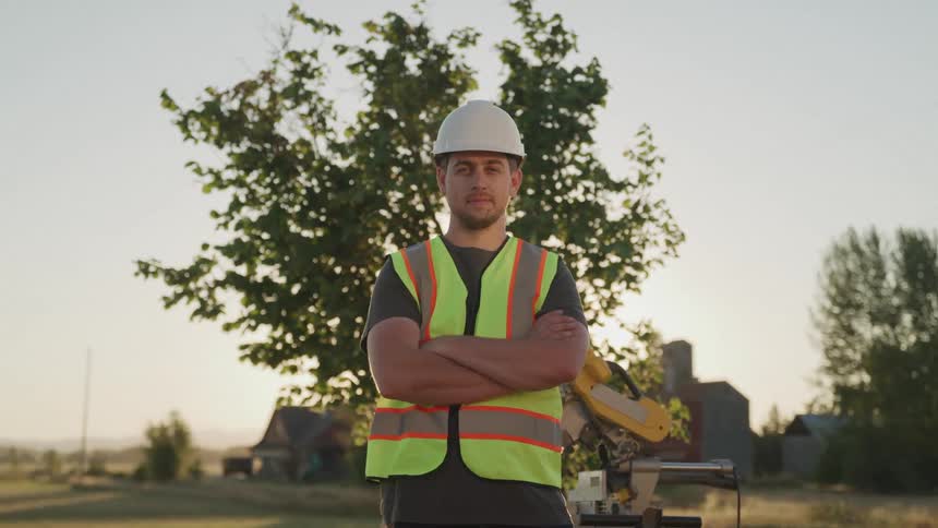Man in safety vest standing in front of tree with arms crossed at construction site