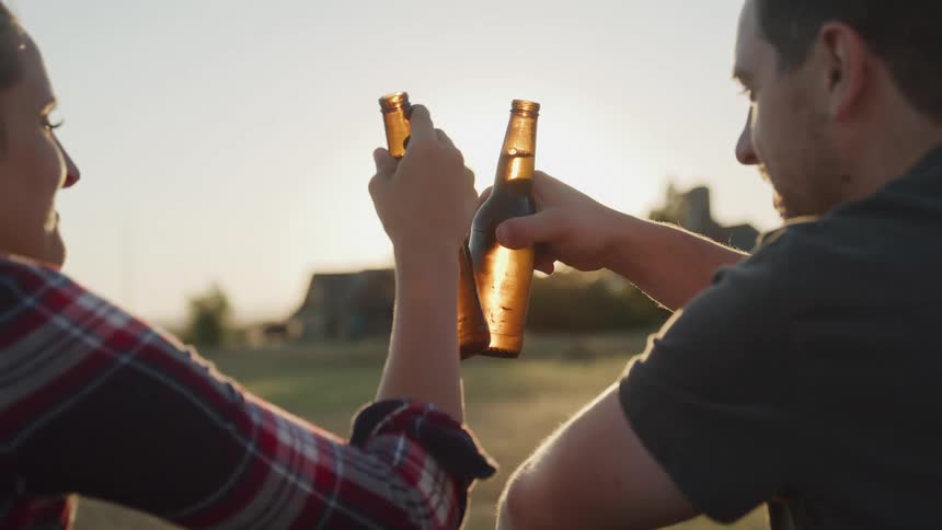 A woman and a man raise glass bottles in a toast with the sun setting in front of them
