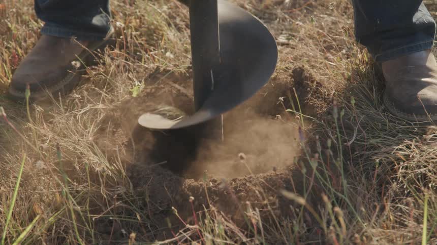 A closeup shot shows a rancher using a mechanical auger to drill a hole into the dry grassy ground as dirt flies upwards