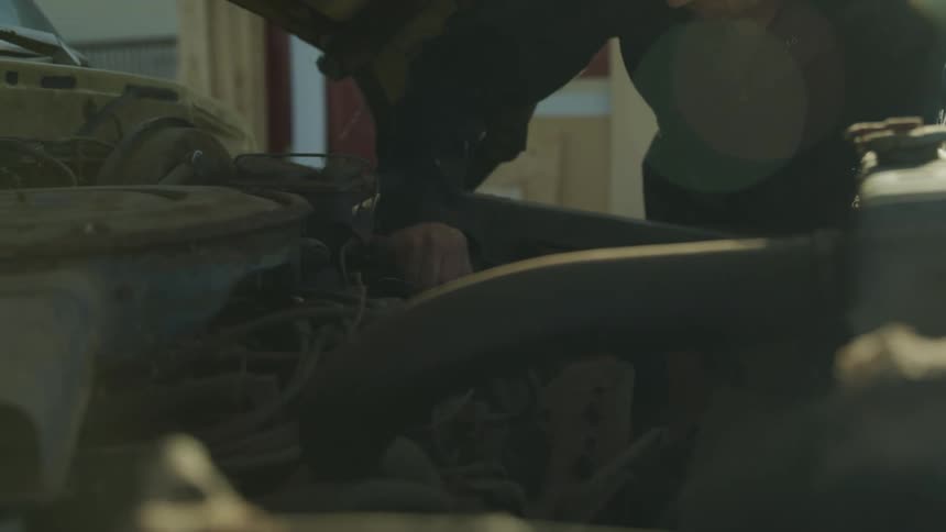 A man in work clothes leans under the hood of a classic car working on the engine