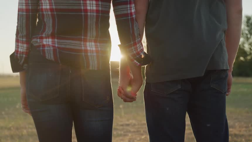 A couple holds hands with sunlight streaming through between them in an open field