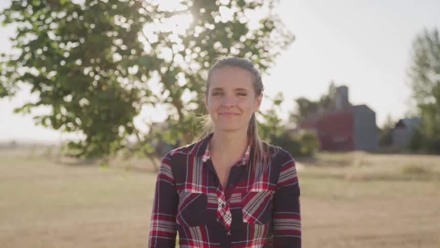 A young woman in a plaid shirt smiles at the camera while standing in a sunny rural field with a tree and farmhouse in the background