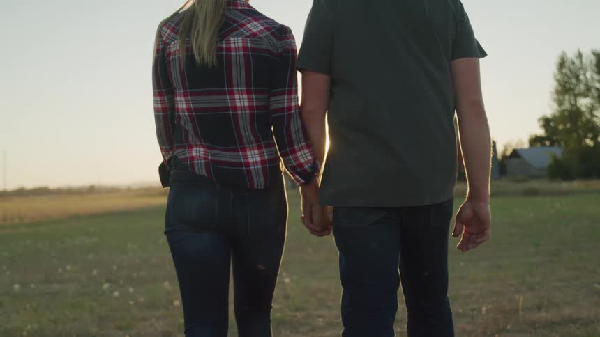 A couple walk hand in hand through a grassy field at sunset with the sun shining brightly between them