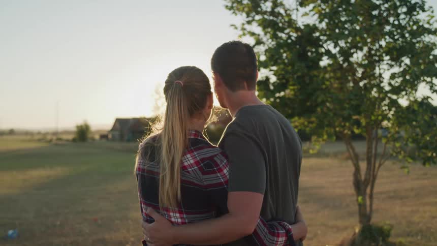 A couple stands side by side outdoors arms around each others waists gazing at the horizon