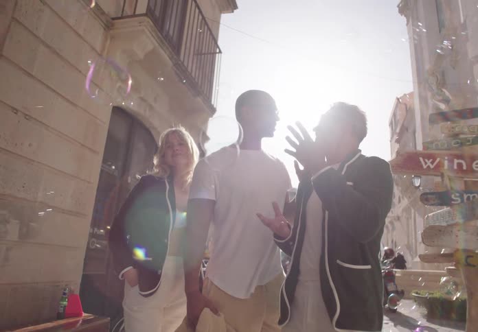 Three people talk on a sunlit street beside a colorful signpost surrounded by floating soap bubbles