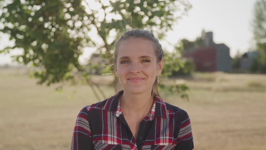 A young woman smiles at the camera while standing in a sunlit rural field