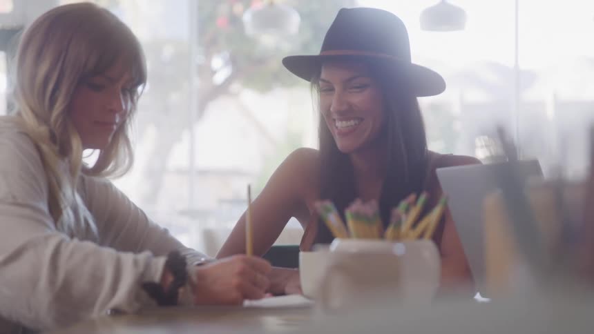Two women sitting at a table with one of them writing are talking