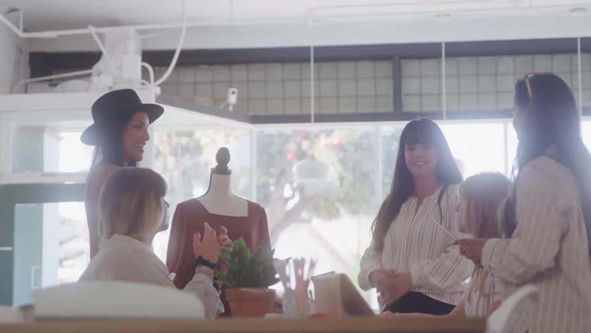 A designer stands beside a mannequin in a sunlit room explaining the displayed dress to a group of women