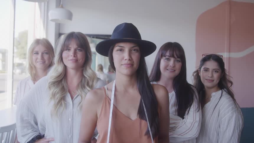 Five women pose confidently in a bright studio led by one in a hat with a measuring tape