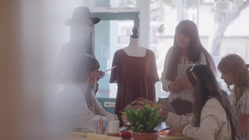 A group of fashion designers collaborate around a table discussing a brown dress on a mannequin