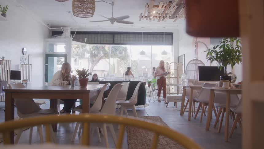 A cozy sunlit cafe shows a few people relaxing and working at wooden tables in a calm airy setting