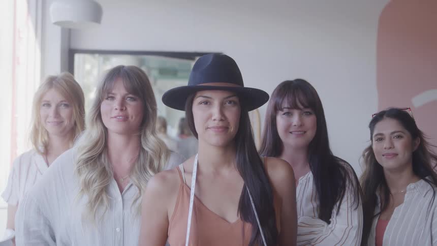Five women stand side by side indoors with smiles on their faces the one in front wears a hat and has a measuring tape draped around her neck around midday