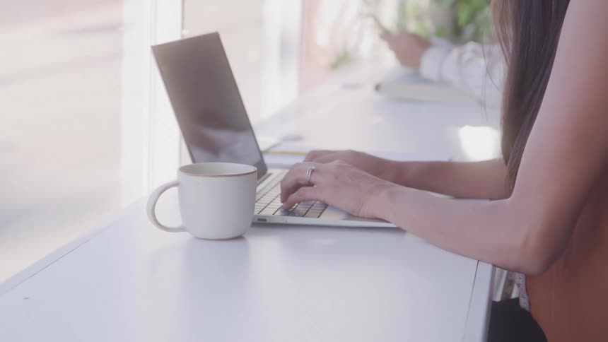 A woman works on a laptop at a bright cafe counter with a coffee mug beside her