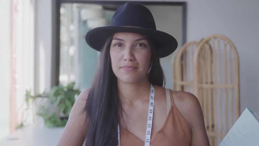 A woman wearing a dark hat and an orange top with a measuring tape around her neck looks forward