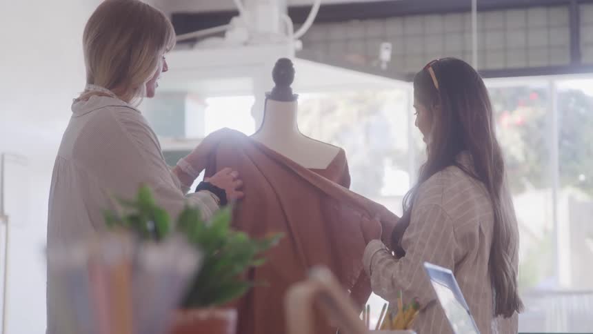 A fashion designer collaborates with her client while draping fabric on a dress form in a bright studio